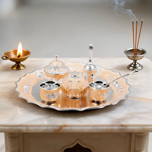 Peach and silver German Silver pooja thali set with bell, bowls, lotus and a lit diya on a marble table